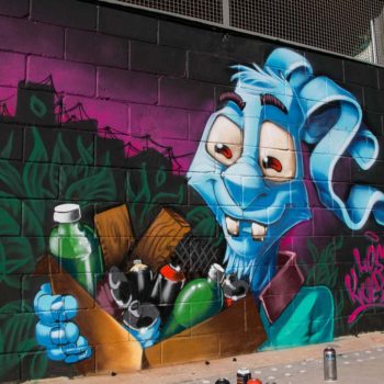 Artista: Kueio. Descrição: Parede externa apresenta grafitti de um coelho azul segurando uma caixa de papelão com diversos resíduos para reciclagem.