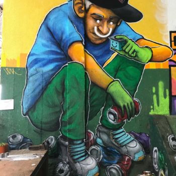 Artistas: ONH. Descrição: Parede interna com fundo amarelo e verde apresenta grafitti de homem sentado recolhendo latinhas e olhando para uma caixa em sua mão.
