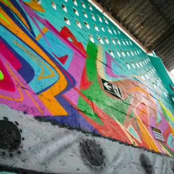 Artista: Sprart. Foto: Júlia Dávila. Descrição: Parede interna interna com grafitti de formas abstratas em diferentes cores como verde, rosa, vermelho e amarelo, azul e verde limão.