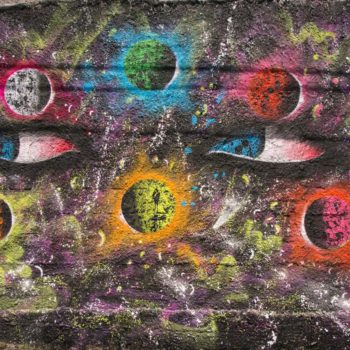 Artista: Saci. Descrição: Muro externo preto apresenta grafitti com diferentes planetas coloridos e no meio deles é possível ver dois olhos.