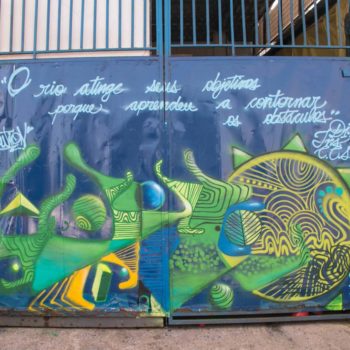 Artista: Evolution. Descrição: Muro externo azul, apresenta grafitti com formas assimétricas nas cores verde amarelo e azul. acima pode-se ler a frase "o rio atinge seus objetivos porque aprendeu a contornar os obstáculos".