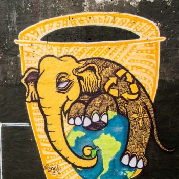 Artista: Lanesky. Descrição: Muro externo preto apresenta grafitti de lata de lixo amarela, na frente dela um elefante amarelo com traços pretos abraça o planeta terra.