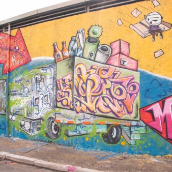 Artista: Alex23 e Bart. Descrição: Muro externo amarelo apresenta grafitti de um caminho de lixo colorido, cheio de material reciclado.