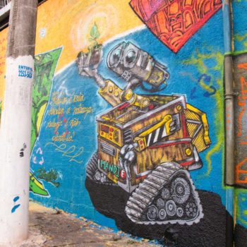 Artista: Mandie. Descrição: Muro externo amarelo apresenta grafitti de um robô construído com partes de lixo eletrônico. o robô está segurando uma planta em um vaso.