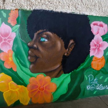 Artista: Pita Isa. Descrição: Parede interna da cooperativa apresenta um mural graffiti de uma mulher negra de cabelos blackpower. A mulher se encontra de perfil. Ela tem olhos verdes claros e está envolta de flores de cores vibrantes (laranja, amarelo e rosa). O fundo da parede é verde.
