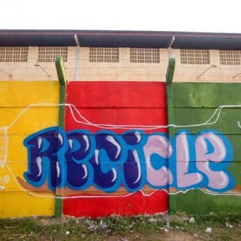 Artista: Prisk. Descrição: Muro externo da cooperativa apresenta mural graffiti de uma garrafa pet deitada. Ela é transparente e dentro se encontra a palavra Recicle.