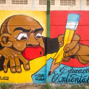 Artista: DK. Descrição: Muro externo da Cooperbrava apresenta mural graffiti de um homem negro de boca vermelha e camiseta preta escrevendo com um lápis amarelo de madeira em um papel azul. Está escrito: Educação ambiental.