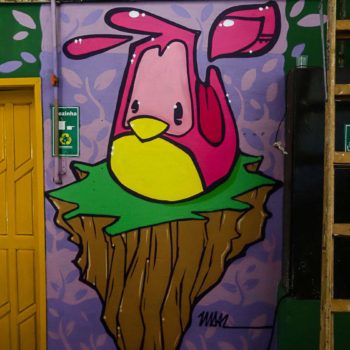 Artista: Vidal. Descrição: Parede interna da cooperativa apresenta mural graffiti de um passarinho de rosa de peito amarelo. O passarinho, de bico curto e amarelo, está sentado em um pedaço de grama verde. O fundo da parede e do desenho apresenta cor roxa clara.