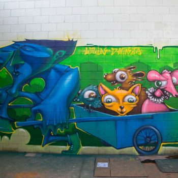 Artista: Lelin e Dninja. Descrição: Parede interna bege apresenta grafitti no fundo colorido verde com personagem azul puxando uma carroça com quatro personagens que se assemelham a animais nas cores laranja, azul, marrom e rosa.