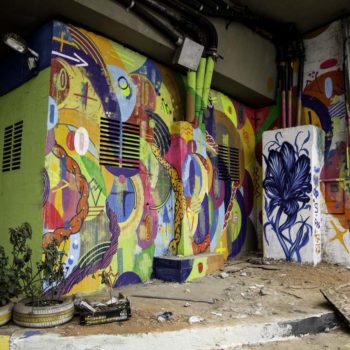 Artistas: Leiga e Mateus Bailon. Descrição: Muro externo de uma cooperativa de recicláveis se encontra pintado de branco. No centro, se encontram dois murais graffiti multicoloridos e sobrepostos, de formas abstratas e geométricas.