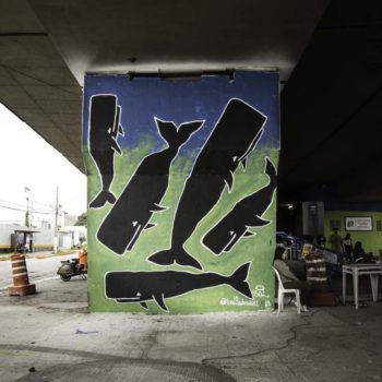 Artista: Bal. Descrição: Coluna externa de um viaduto apresenta mural graffiti de fundo azul e verde vibrantes. Sobre a pintura, estão desenhadas 5 baleias pretas.
