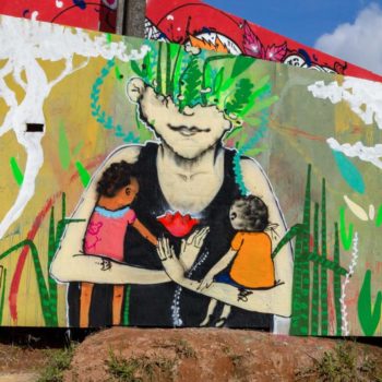 Artista: Ayco Dany. Descrição: parede externa com fundo verde apresenta arte graffiti com mulher vestida de preto abraçando duas crianças. Ao redor da mulher com as duas crianças, se encontram plantas, como espada de são jorge. No canto inferior direito se encontra um gato preto.