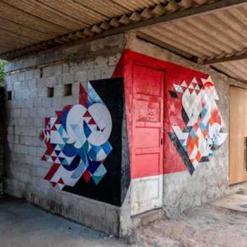 Artistas: João e Diego, do coletivo Muda. Descrição: parede de cimento que apresenta um mural artístico abstrato. O mural é produzido com pedaços de azulejos coloridos cortados que formam um mosaico. Ao fundo, a parede está pintada de preto e vermelho.
