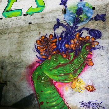 Artista: Frenesi. Foto Júlia Dávila. Descrição: Parede interna branca apresenta grafitti de uma mão verde, segurando diversas flores em roxo e laranja. No meio das fllores está o desenho da terra.