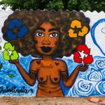 Artista: Thainá India. Foto: Júlia Dávila. Descrição: Muro externo branco apresenta grafitti de mulher índia nua segurando nas mãoes os símbolos de reciclagem nas cores verde, vermelho, azul e laranja. No seu colo está escrito “recicle”.