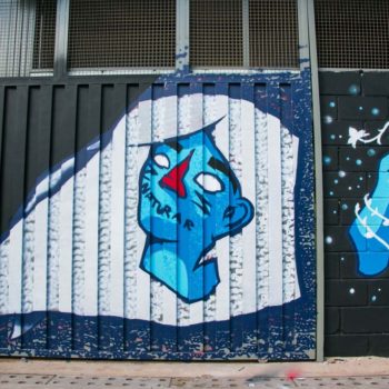 Artista: El Doze. Descrição: Parede externa com fundo preto e azul apresenta grafitti de rosto azul com longos cabelos brancos. ao lado uma mão azul segura uma planta crescendo.