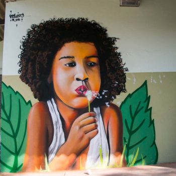 Artista: Krusty. Descrição: parede externa bege apresenta grafitti de rosto de uma menina negra assoprando uma flor.