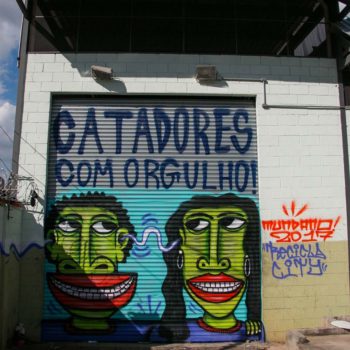 Artista: Mundano. Descrição: Portão externo com fundo azul apresenta grafitti dos rostos verdes de um homem e uma mulher. acima deles lê-se a frase "catadores com orgulho".