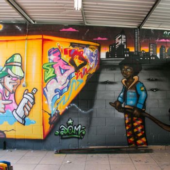 Artista: Dom. Descrição: parede interna apresenta grafitti de um muro amarelo com diversos grafitittis e enquanto um rapaz negro puxando uma carroça de reciclagem o observa.