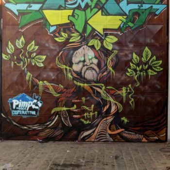 Artista: OTITO e Mandi. Foto: Arthur Moriyama. Descrição: Portão externo de fundo marrom apresenta grafitti de uma árvore, o seu tronco é formando pelo rosto de um homem branco e velho, como um ancião. A copa da árvore forma o símbolo da reciclagem com as cores verde, amarelo e azul.