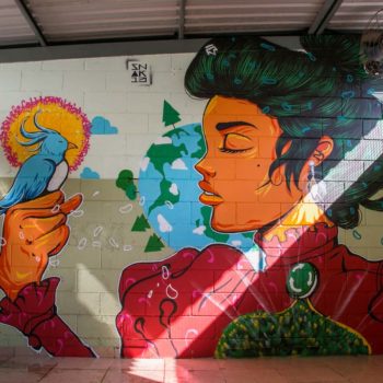 Artista: Snak 13. Descrição: Parede interna bege e branca apresenta grafitti com mulher de pele laranja, vestido vermelho e grandes cabelos presos segura pássaro azul com a mão direita