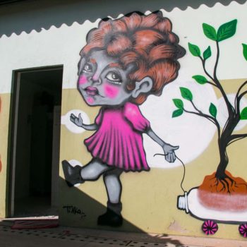 Artista: Tikka. Descrição: Parede externa bege apresenta grafitti de uma menina de pele cinza com vestido rosa puxando um carrinho feito de garaffa pet transparente e dentro dele um pé de árvore.