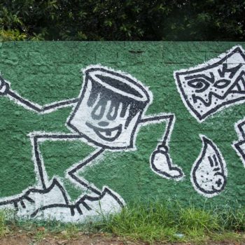 Artista: Felipe Risada. Foto: Rebeca Figueiredo. Descrição: muro externo verde com grafitti de uma lata de tinta com pernas longas e ao lado um rolo de pintura em preto e branco.