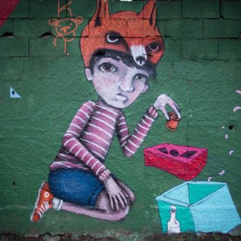 Artista: Kot. Foto: Rebeca Figueiredo. Descrição: muro externo verde com grafitti de uma menina sentada usando um gorro com formato da cabeça de uma raposa marrom. ela brinca com cubos na cor verde e vermelho.