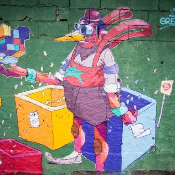 Artista: Grego. Foto: Rebeca Figueiredo. Descrição: muro externo verde com grafitti de uma forma humana com cabeça de pássaro vestindo um macacão roxo cercado por latas de lixo nas cores vermelho, amarelo e azul. na sua mão uma construção pequena de blocos coloridos.