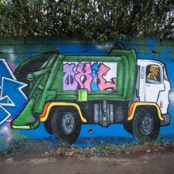 Artista: Oxil. Foto: Rebeca Figueiredo. Descrição: muro externo azul com grafitti de um caminhão de lixo colorido, saindo da traseira dele o símbolo de reciclagem em azul. ao centro escrito oxil em branco.