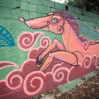 Artista: 3visão. Foto: Rebeca Figueiredo. Descrição: muro externo verde com grafitti de figura semelhante a um cavalo na cor rosa, em meio a nuvens vermelho escuro contornadas de rosa.