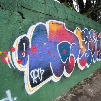 Artista: Gueto. Foto: Rebeca Figueiredo. Descrição: muro externo verde com grafitti de letras diferentes nas cores roxa e amarela, com contorno branco.