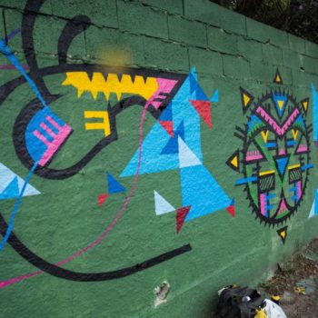 Artista: Márcio Ficko. Foto: Rebeca Figueiredo. Descrição: muro externo verde com grafitis de triângulos coloridos em diferentes tons de azul e vermelho.