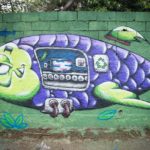 Artista: Gui Matsumoto. Foto: Rebeca Figueiredo. Descrição: muro externo verde com grafitti de um peixe robótico nas cores roxo e verde. dentro dele é possível ver uma esteira por onde correm caixas.