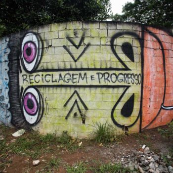 Artista: Mundano. Foto: Rebeca Figueiredo. Descrição: muro externo cinza com grafitti de um rosto verde na horizontal, com olhos roxos e lábios grandes vermelhos. no seu rosto está escrito “reciclagem e progresso”.