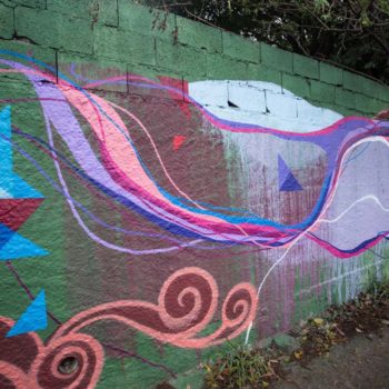 Artista: Ricardo Célio e Ficko. Foto: Rebeca Figueiredo. Descrição: muro externo verde com grafitti de formas abstratas cor de rosa e azul claro. ao lado triângulos azuis e vermelhos