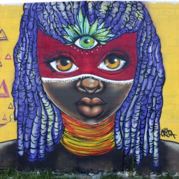 Artista: Crica. Descrição: Parede com fundo amarelo apresenta o grafitti de uma mulher africana com uma máscara vermelha deixando descoberta seus olhos cor de mel. Seu cabelo é lilás e no meio de seus olhos um destaque para o terceiro olho na cor verde.