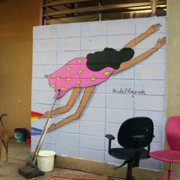 Artista: Paula P. Rezende. Descrição: parede interna com fundo azul claro, apresenta grafitti de uma mulher de costas voando com uma capa cor de rosa estampada com folhas verdes.