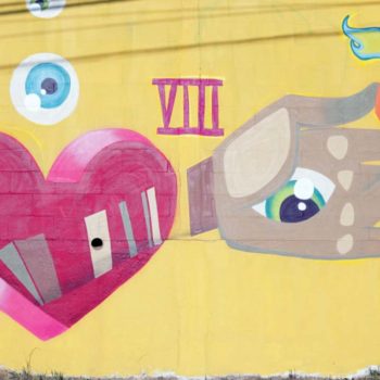 Artista: Elisa Fresz. Descrição: Muro externo com fundo amarelo, apresenta grafitti com coração vermelho saindo dele uma mão segura um fósforo acesso.