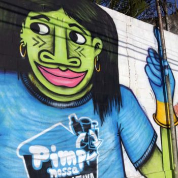 Artista: Mundano. Descrição: Muro externo com fundo branco, apresenta grafitti com corpo de uma mulher, seu rosto é verde, vestindo camiseta com o logo do Pimp Nossa Cooperativa onde predomina o azul claro e contornos na cor branca.