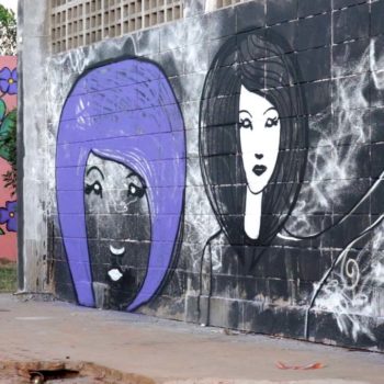 Artista: Mathiza. Descrição: Muro externo natural, na cor preta, apresenta grafitti com desenho do contorno de dois rostos femininos, o primeiro com o cabelo roxo, o segundo com cabelo preto.