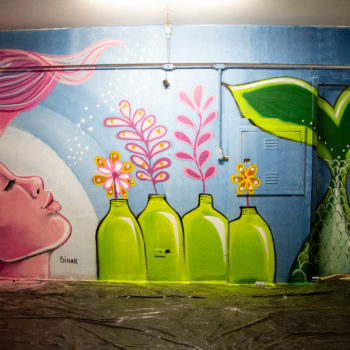 Artista: Binak. Descrição: Parede interna da Cooperativa Central Tietê apresenta mural artístico de uma sereia de rosto rosa claro. A sereia apresenta feição serena e uma longa cauda verde. No desenho, algumas garrafas simbolizam materiais reciclados.