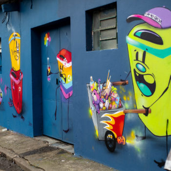 Artista: Feik. Descrição: Parede externa da Cooperativa Central Tietê apresenta mural artístico de latas de lixo humanizadas. As lixeiras são coloridas e apresentam olhos e boca. Uma delas carrega um carrinho com materiais reciclados.