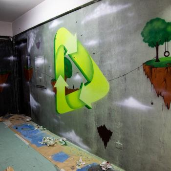 Artista: Nossa. Descrição: Parede interna da Cooperativa Central Tietê apresenta mural artístico do símbolo de materiais recicláveis. Ao lado, pequenos pedaços de terra, com árvores plantadas flutuam no ar.