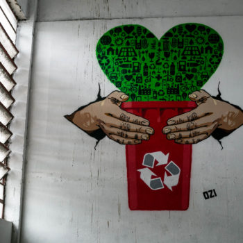 Artista: Ozi. Descrição: Parede interna da Cooperativa Central Tietê apresenta mural artístico em stêncil. Duas mãos seguram uma lata de lixo vermelha. Da lata, surge um coração verde com símbolos de sustentabilidade.