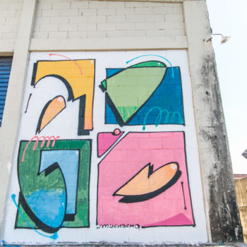Artista: Muchacho. Descrição: Em um paredão com aproximadamente de 4 metros de altura, um grafitti com quatro quadrados, dois em cima e dois embaixo, nas cores amarelo, branco, azul, verde e rosa. Em cada um, uma figura abstrata tridimensional, ocupa parte do quadrado.