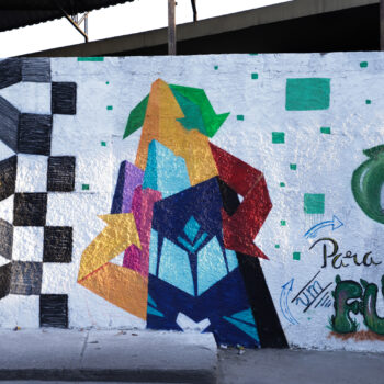 Artista: Wagner Maciel | Descrição: Sobre muro branco e crespo, um desenho geométrico lembra um conglomerado de prédios com o topo triangular e sobreposições de retângulos e na vertical, um azul, um rosa, um lilás e o da base tem algumas figuras geométricas quadriláteras. O símbolo da reciclagem envolve a estrutura geométrica. No alto, uma seta verde aponta para baixo, na lateral direita, uma vermelha e na lateral esquerda, uma amarela aponta para cima.