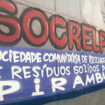 Artista: Mundano | Descrição: Grafite em portão de chapas de metal cinza. Em letras vermelhas com contorno preto: SOCRELP. Em letras menores brancas sobre faixa azul: Sociedade comunitária de reciclagem de resíduos sólidos do Pirambu.