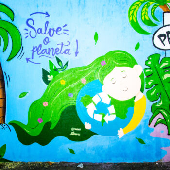 Artista: Manu. Descrição: Graffiti em um muro, de uma garota abraçada ao planeta Terra. Ela tem cabelos verdes, longos e ondulados, decorados com flores coloridas. Repousa o rosto com os olhos fechados no planeta. Usa camisa de manga comprida amarela. No meio do planeta, o símbolo de reciclagem em branco. Mais acima, em letras azuis, “Salve o planeta”. À esquerda, um coqueiro e parte de um grafismo em verde. E à direita, parte do graffiti de um ser com rosto de galinha e corpo humano. Próximo ao bico dele, em um balão de fala, em letras pretas, “Pru!”.