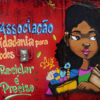Artista: Nega Suh | Descrição: graffiti. Uma garota negra abraçada a um balde com material reciclável e a um vaso com uma planta. Ela olha de lado enquanto arqueia as sobrancelhas. Tem cabelos escuros e cacheados, nariz e boca pequenos. Usa camisa roxa com uma faixa verde. À esquerda, em letras azuis, laranjas e verdes: “Associação Cidadania para todos, Reciclar é Preciso”.